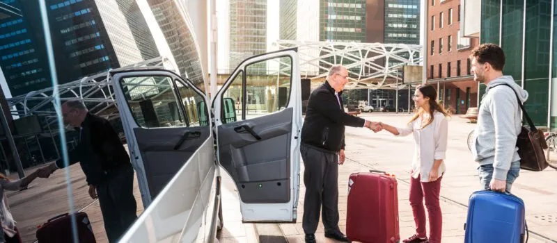 Driver of a airport shuttle minivan, greeting his passengers with their luggage on the sidewalk of a modern city business district